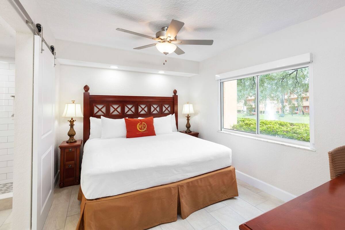 A hotel room with a bed, a window with a view of greenery, a ceiling fan, and a nightstand with a lamp.
