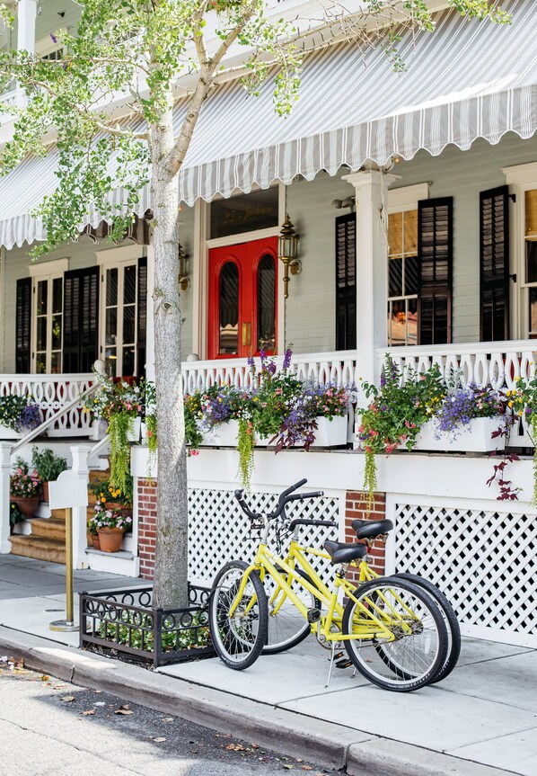 Un vélo jaune est garé sur un trottoir, à côté d’un bâtiment entouré d’une clôture blanche en piquets et agrémenté de jardinières suspendues.