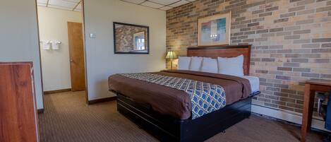 A hotel room with a brick-patterned wall, a bed with a patterned bedspread, a wooden headboard, a nightstand with a lamp, and a desk with a chair.