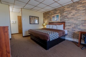 Una habitación de hotel con pared de ladrillo, una cama con colcha estampada, cabecero de madera, mesita de noche con lámpara y un escritorio con silla.