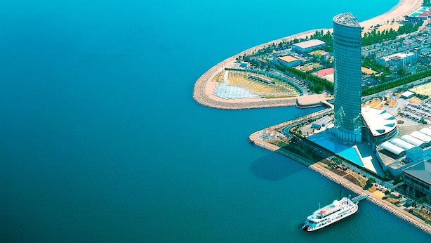 An aerial view of a coastal city with a prominent skyscraper, a marina, and a bridge in the background.