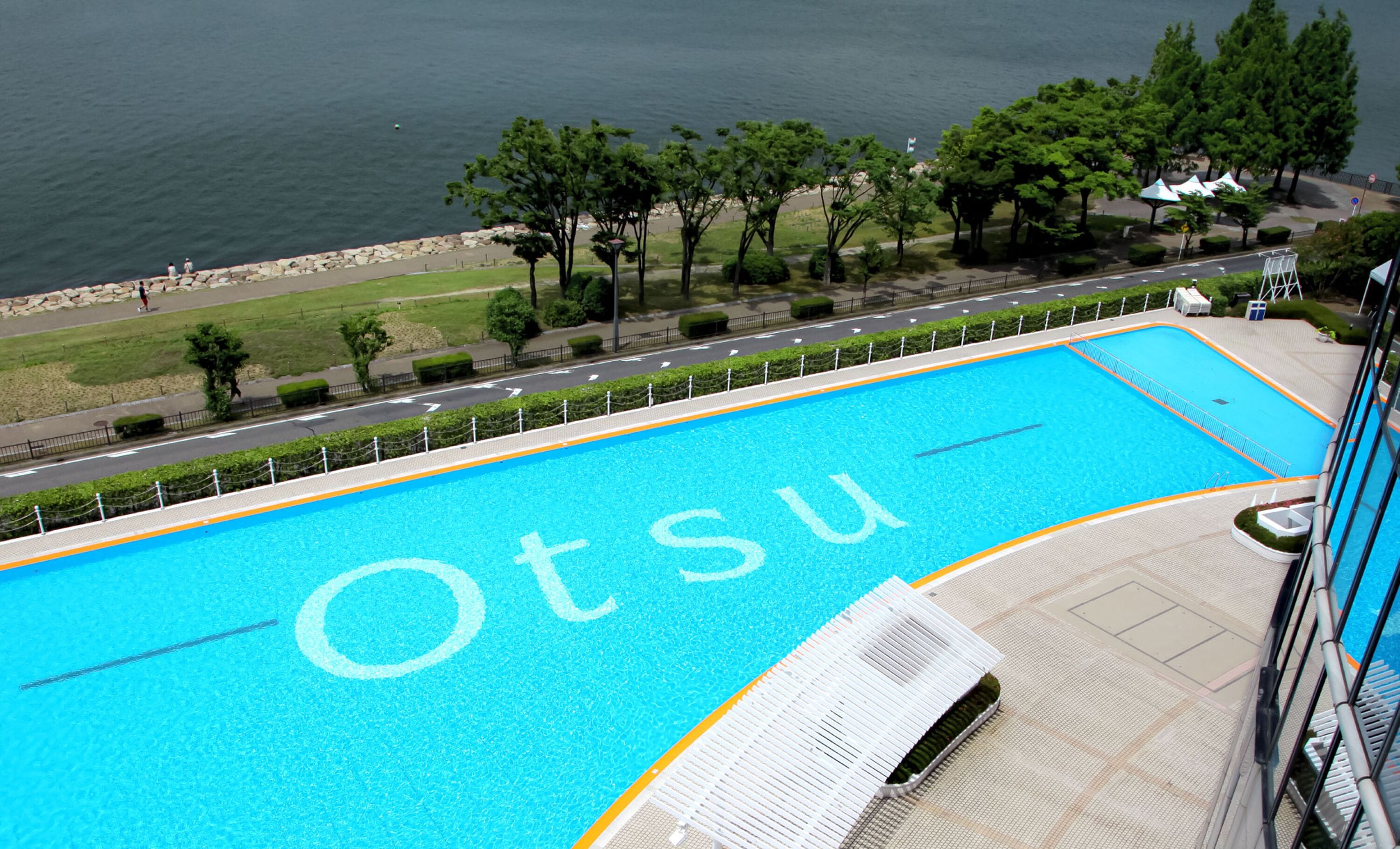 A swimming pool with the name 'Otsu' on the water, surrounded by a paved deck and a view of a road and greenery.