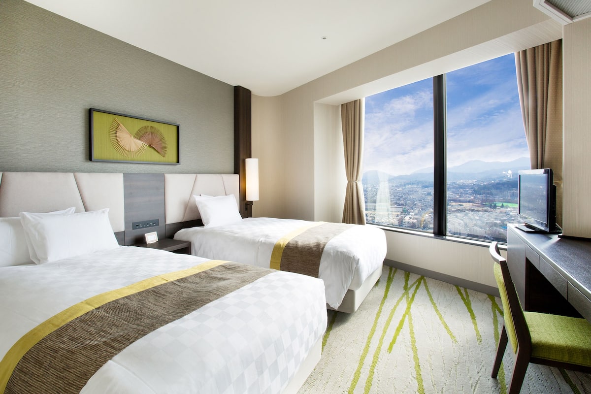 A hotel room with two beds, a desk, a TV, and a large window with a city view.