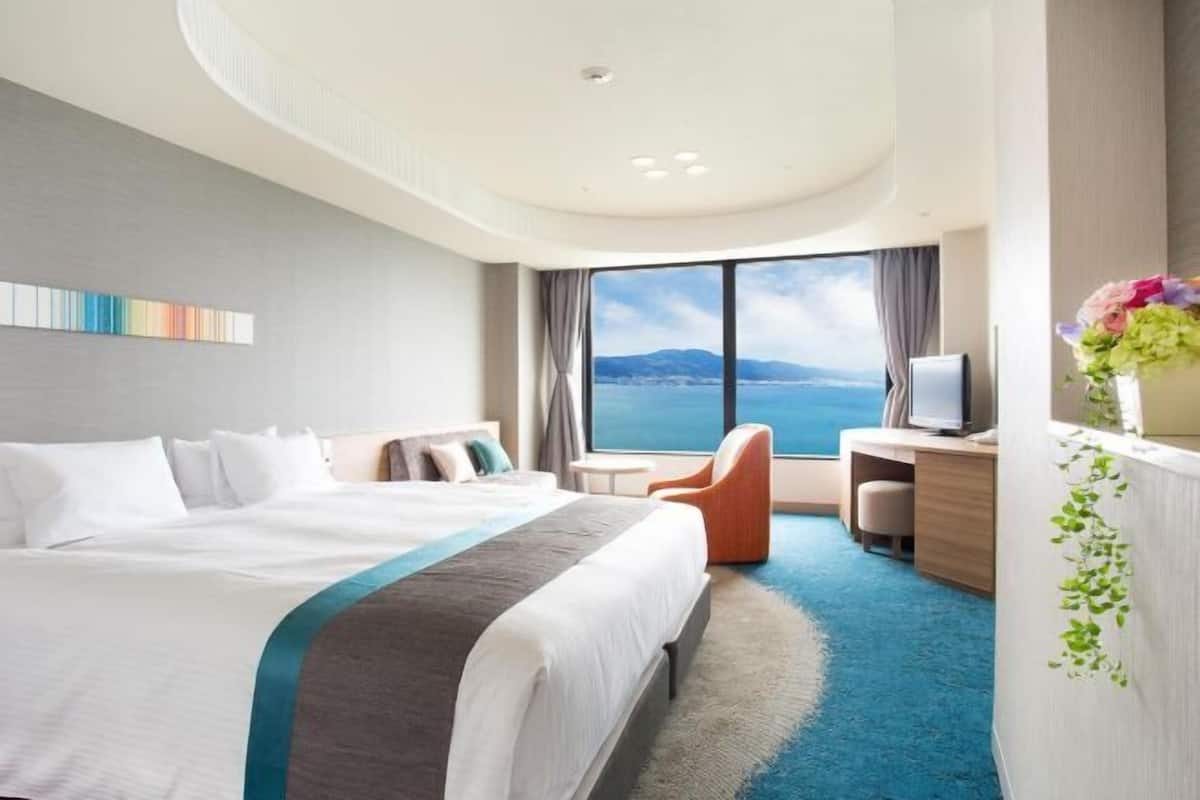 A hotel room with a large bed, a desk, a chair, and a view of the sea.
