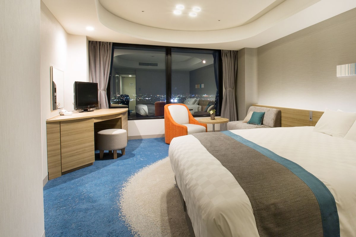 A hotel room with a large bed, a desk, a chair, and a view of the city at night.