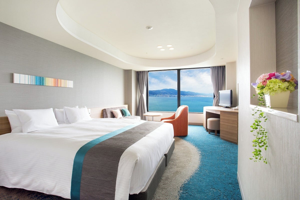 A modern hotel room with a large bed, a desk, a chair, and a view of the sea.