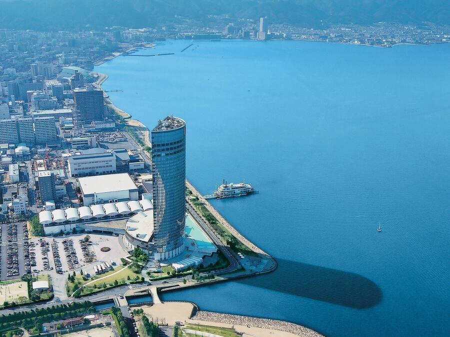 Un edificio alto con fachada curva cerca de la costa, rodeado por un estacionamiento y un área ajardinada.