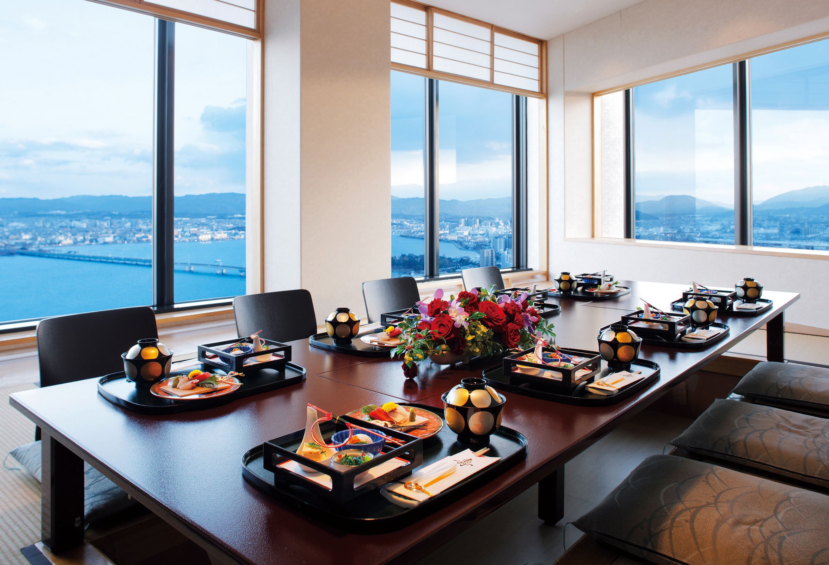 Un salón de conferencias con una larga mesa dispuesta para una reunión, amplias ventanas con vistas a la ciudad y un jarrón con flores rojas.