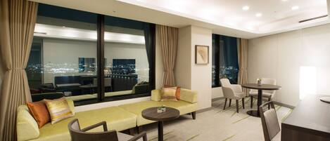 A modern hotel room with a sofa, chairs, a dining table, and large windows offering a city view.