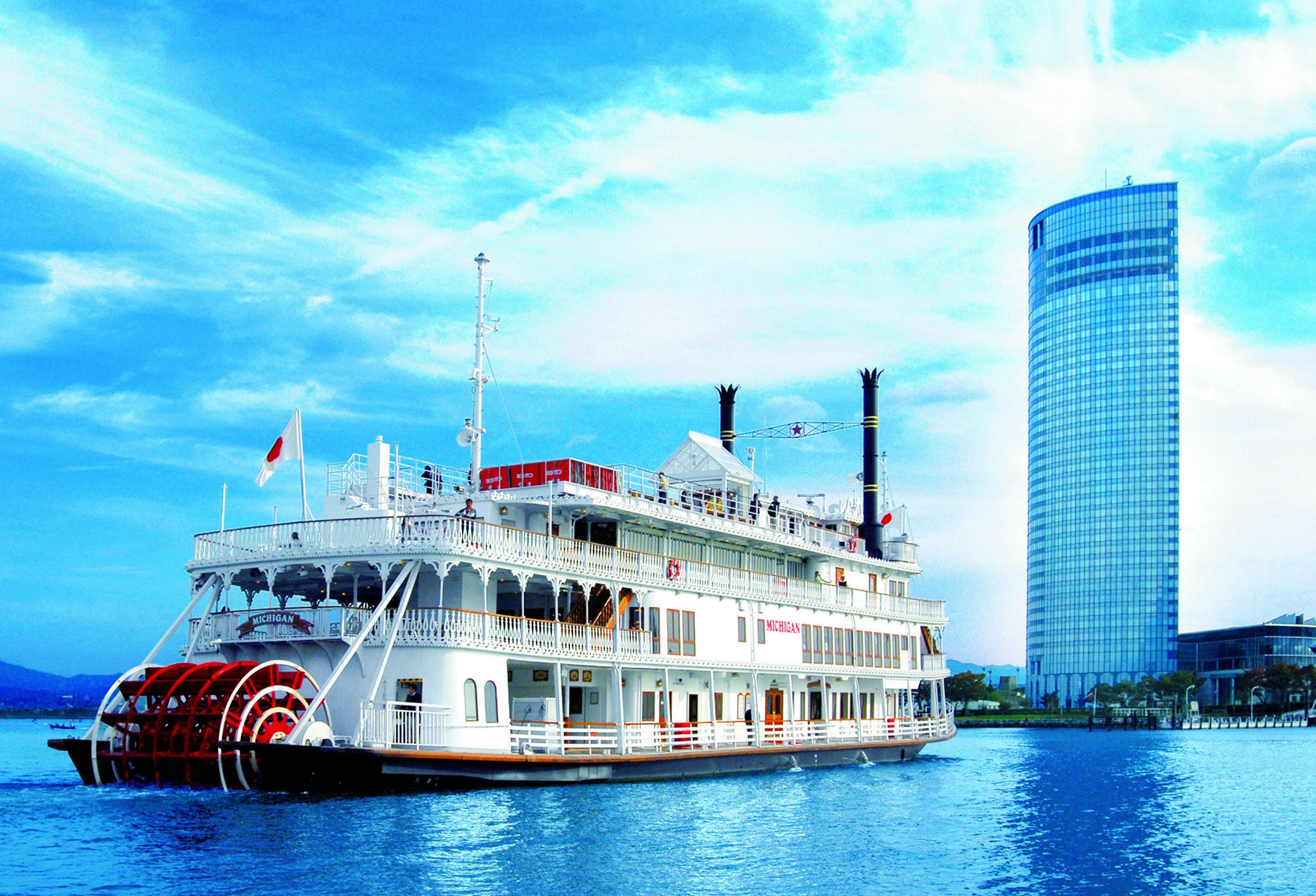 A large riverboat with multiple decks and a flag, docked near a modern skyscraper.