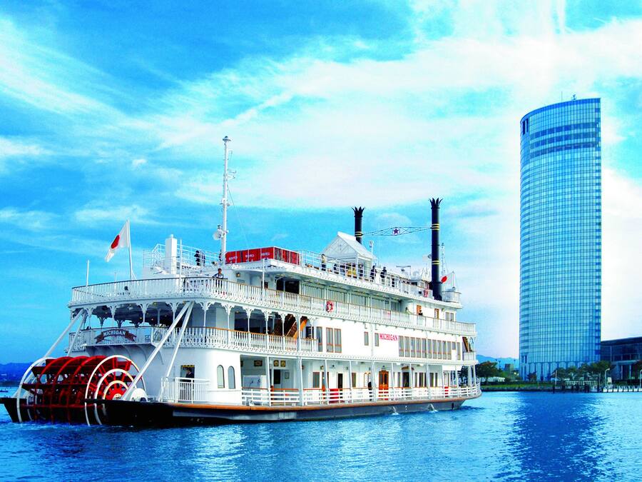 A large riverboat with multiple decks and a flag, docked near a modern skyscraper.