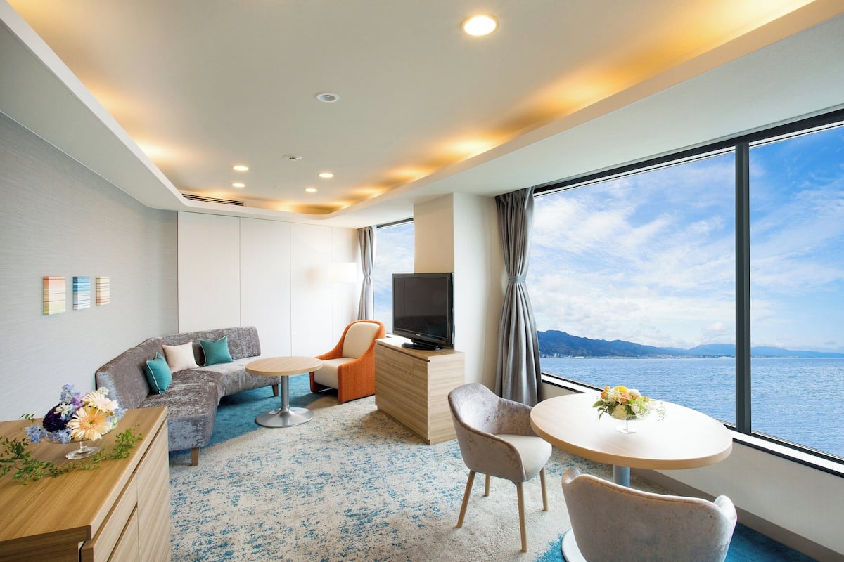 A modern hotel room with a sofa, armchairs, a TV, and a large window offering a scenic view.