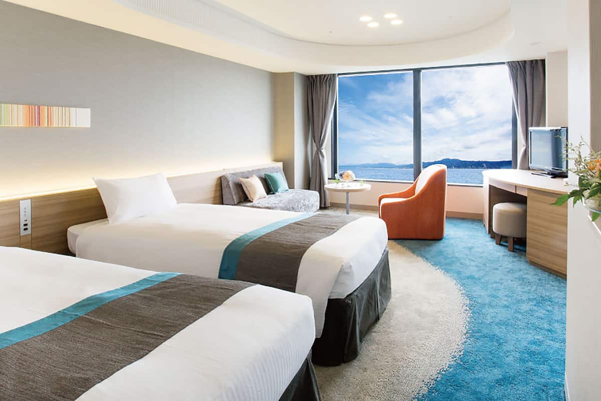 A hotel room with two beds, a desk, a chair, a small table, and a large window offering a view.