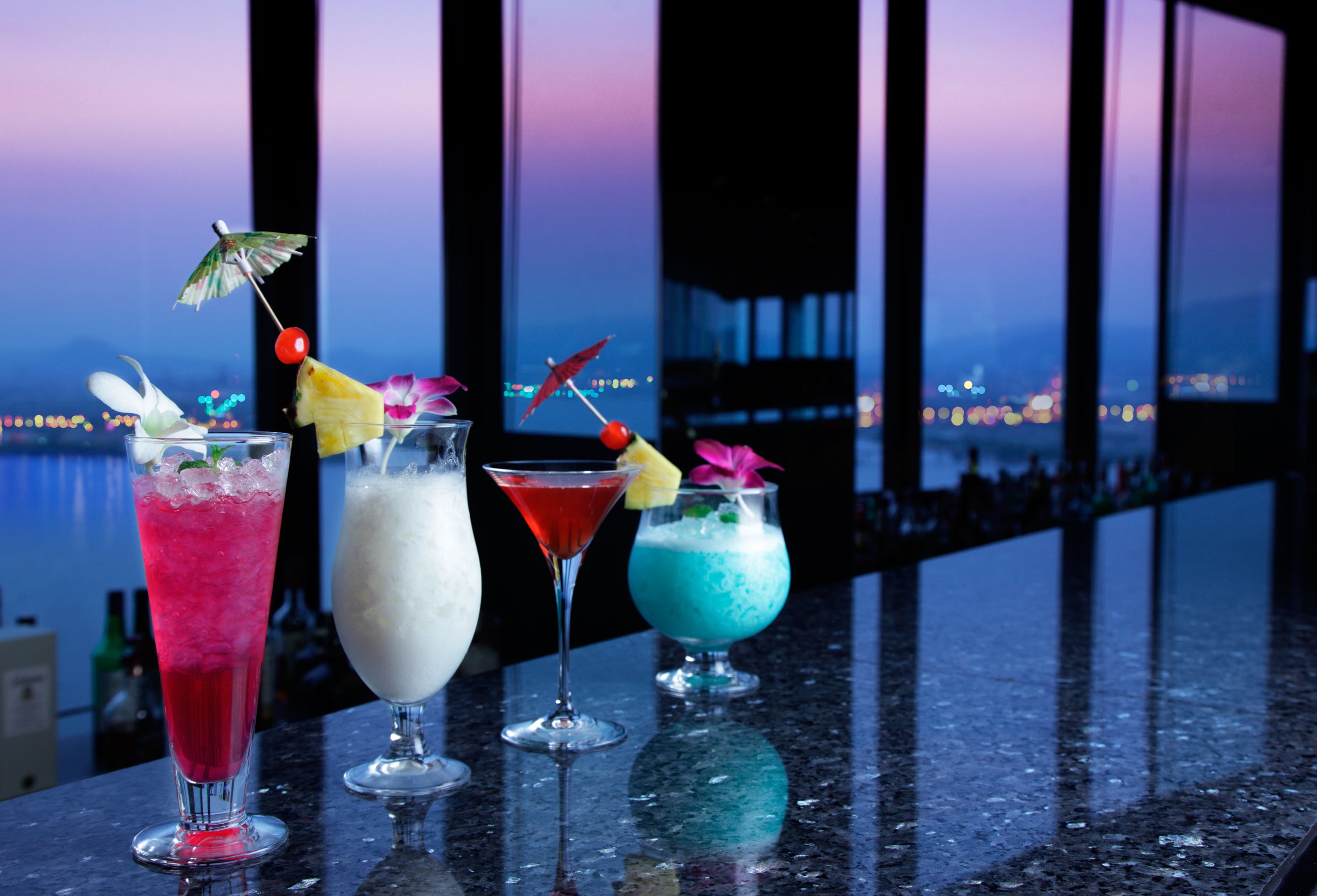 A bar with four colorful cocktails, each garnished with a unique decoration, set against a backdrop of a cityscape at dusk.