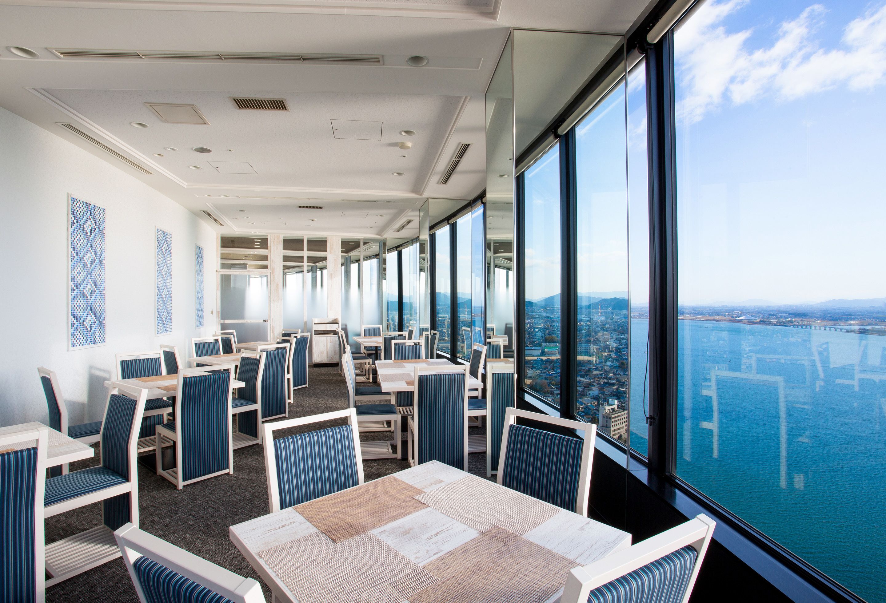 A modern dining area with tables and chairs, large windows offering a city and water view.