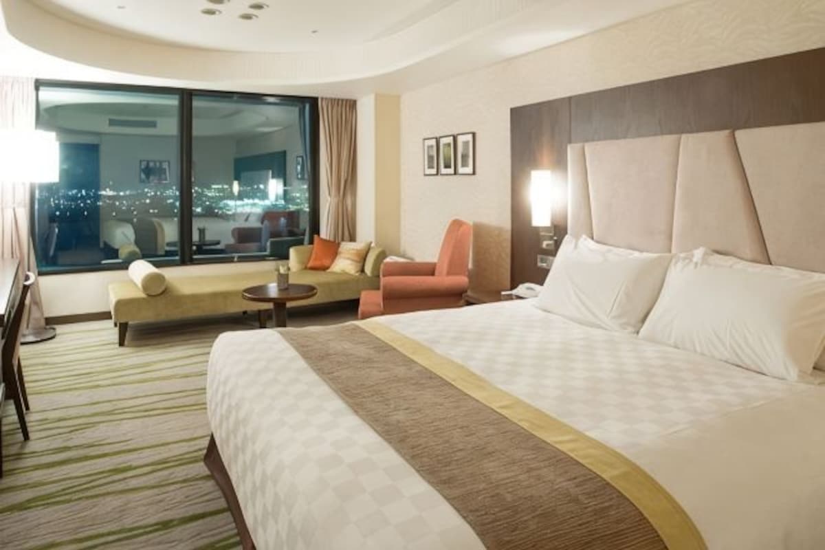 Hotelkamer met een groot bed, een bank, een stoel, een tafeltje en een groot raam met uitzicht op de stad.