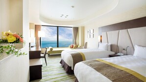 A hotel room with two beds, a desk, a chair, and a large window with a view of the ocean.