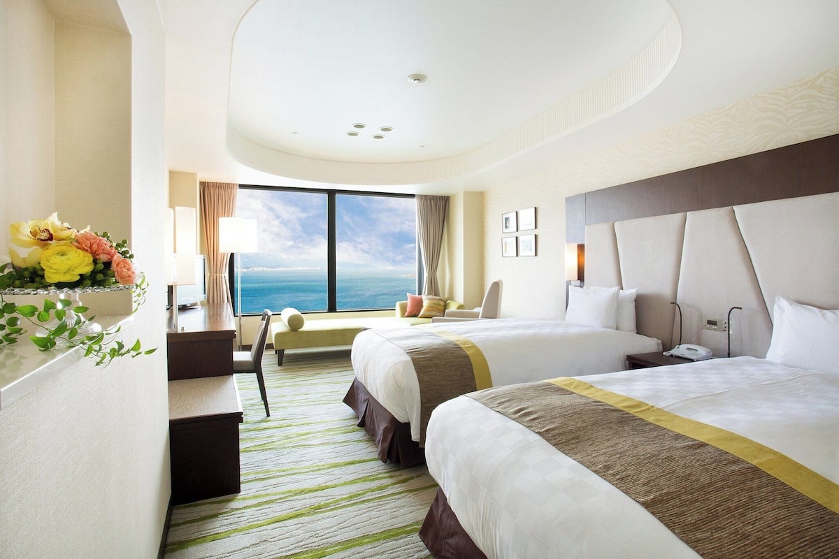 A hotel room with two beds, a desk, a chair, and a large window with a view of the ocean.