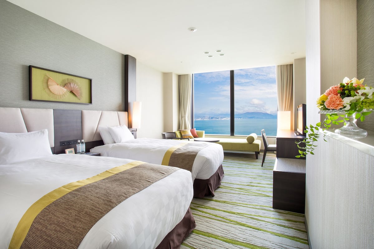 A hotel room with two beds, a large window with a view, a desk, and a plant.