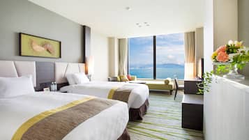 A hotel room with two beds, a large window with a view, a desk, and a plant.