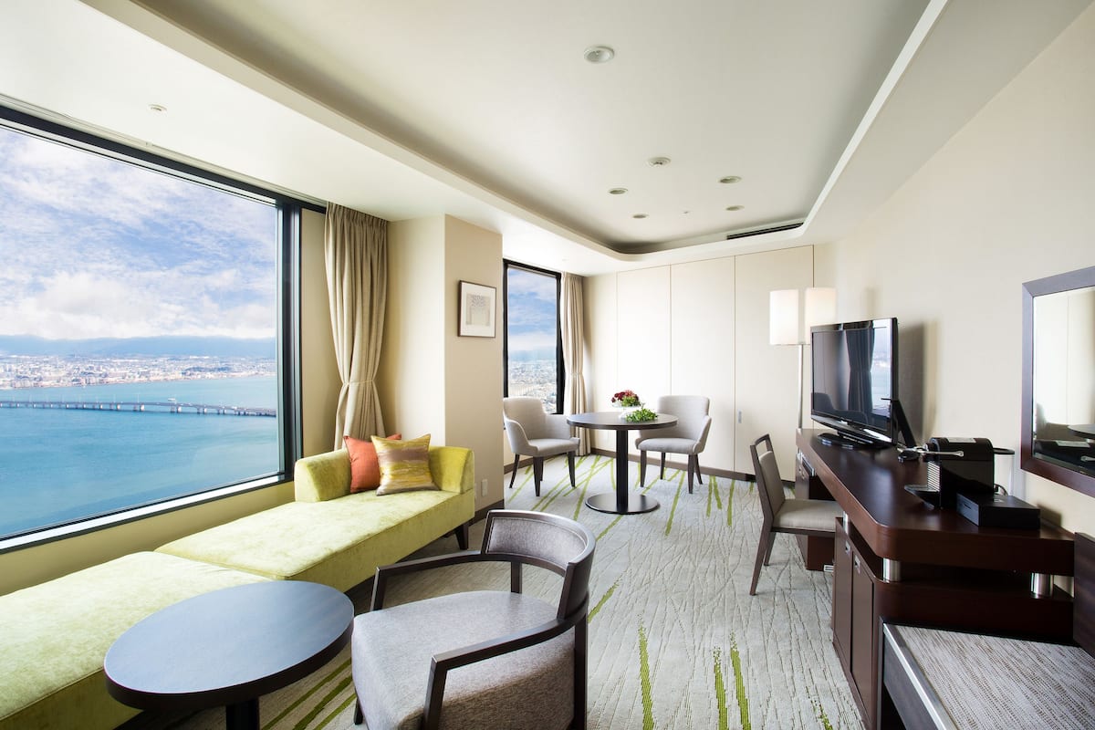 A hotel room with a large window offering a city view, a sofa, a dining area with chairs, a TV, and a small table.