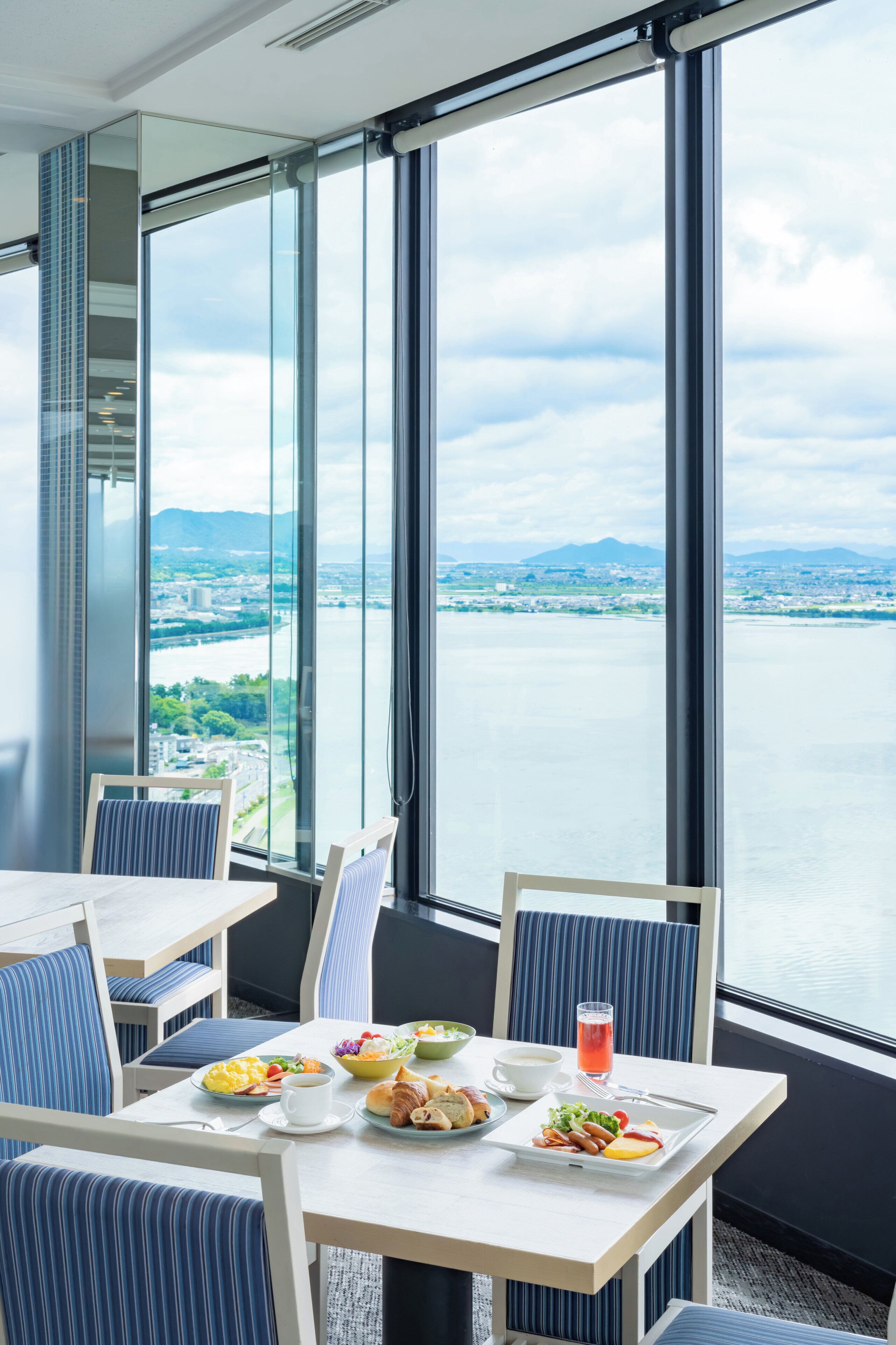 Comedor con una mesa dispuesta para el desayuno, amplias ventanas que ofrecen vistas a un río y montañas a lo lejos.