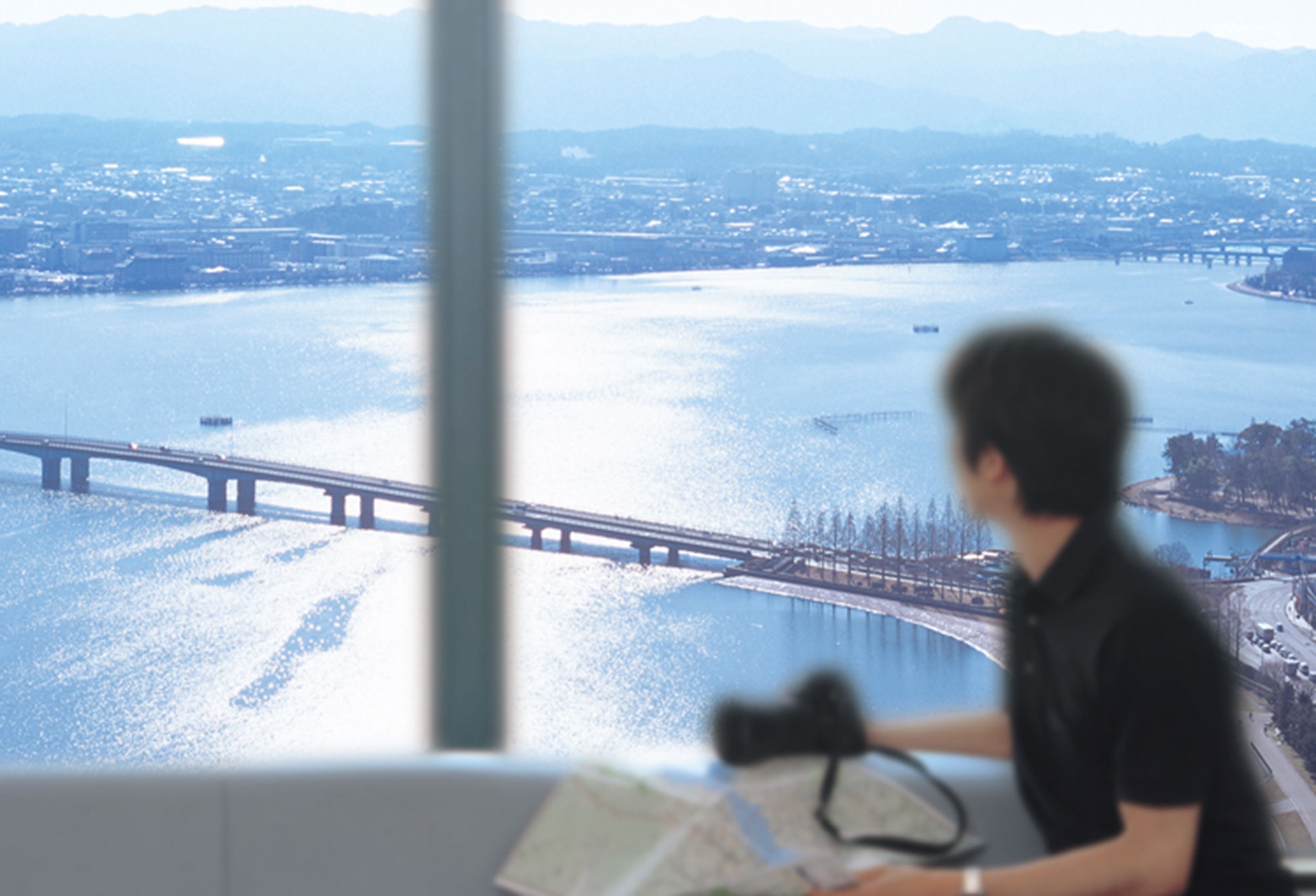 A person sitting by a window overlooking a river with a bridge, with a map and camera on the table.