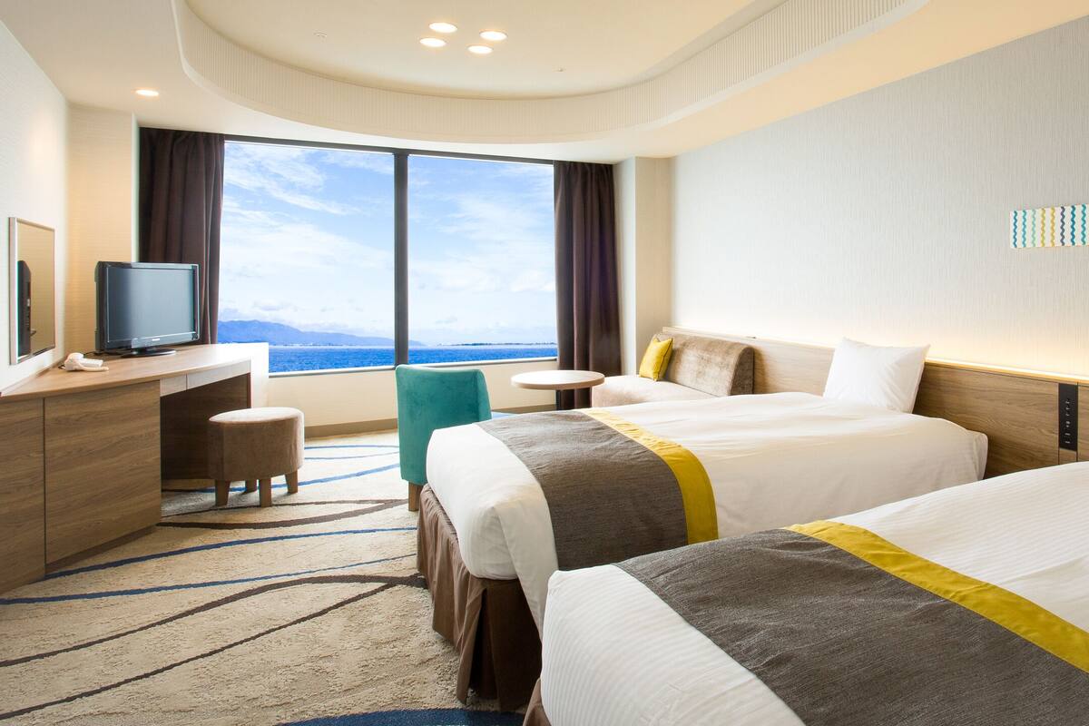 A hotel room with two beds, a desk, a television, and a large window offering a view of the sea.