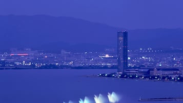 A cityscape at dusk with a prominent skyscraper and a waterfront with fountains.