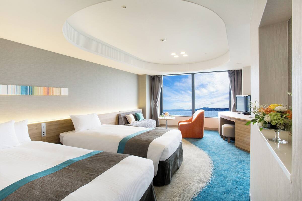 A hotel room with two beds, a desk, a chair, a TV, and a large window with a view.