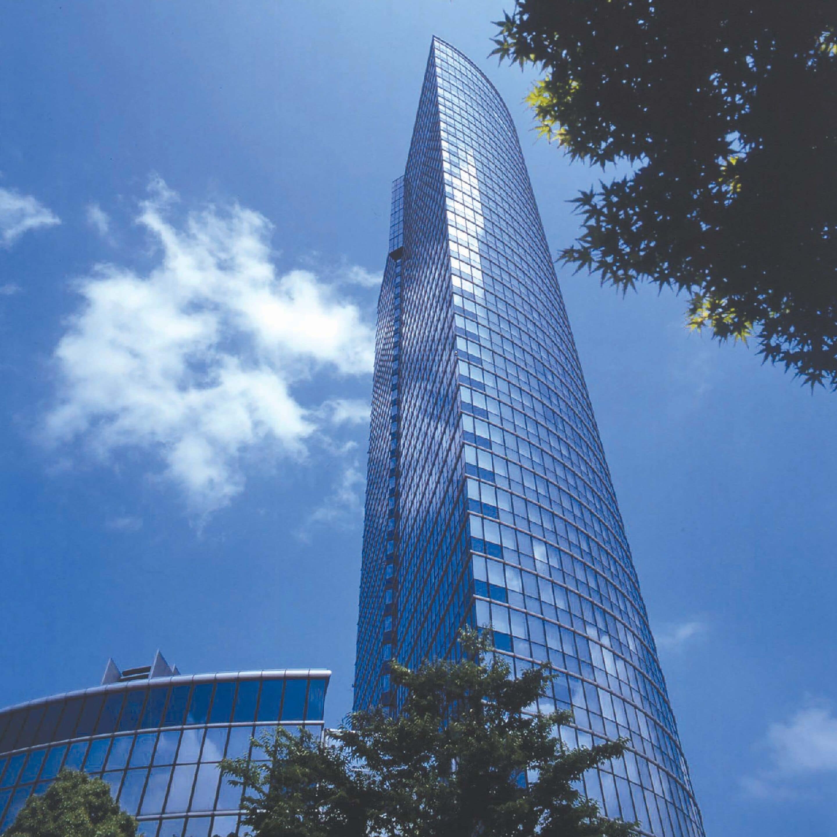 A tall, modern skyscraper with a curved glass facade, surrounded by trees and a clear blue sky.