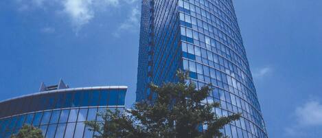 A tall, modern skyscraper with a curved glass facade, surrounded by trees and a clear blue sky.