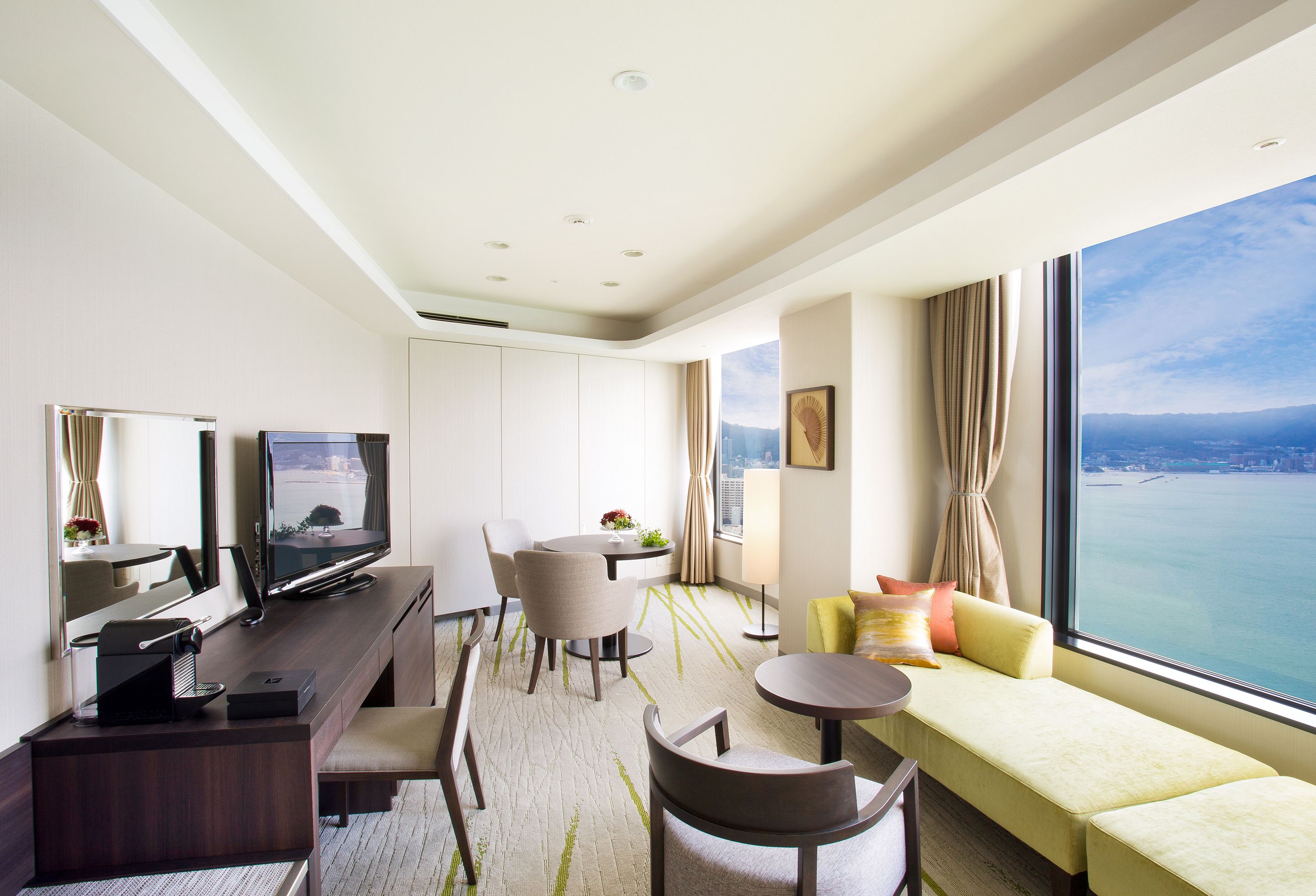 A modern hotel room with a large window offering a city view, a flat-screen TV, a dining area, and a seating area with a sofa and chairs.