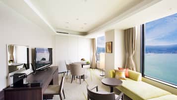 A modern hotel room with a large window offering a city view, a flat-screen TV, a dining area, and a seating area with a sofa and chairs.