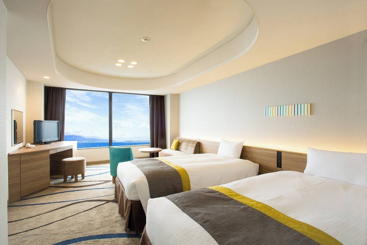 A hotel room with two beds, a desk with a computer, a small stool, a round table, and a large window with a view of the sky and mountains.