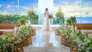 A wedding ceremony with a bride in a white dress, aisle lined with floral arrangements and candles, and a view of the sea through large windows.