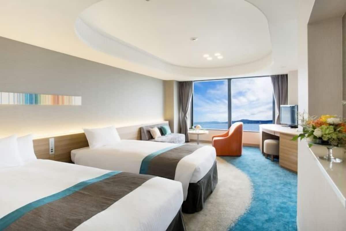 A hotel room with two beds, a desk, a chair, and a large window with a view.