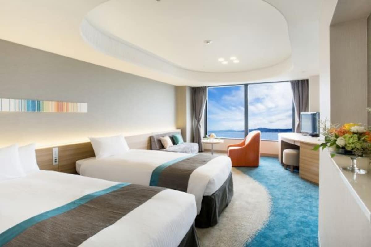 Een hotelkamer met twee bedden, een bureau, een stoel en een groot raam met uitzicht.