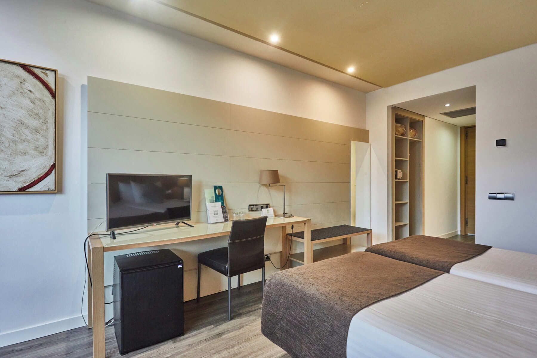 Habitación de hotel con cama, escritorio, computadora, televisor y refrigerador.