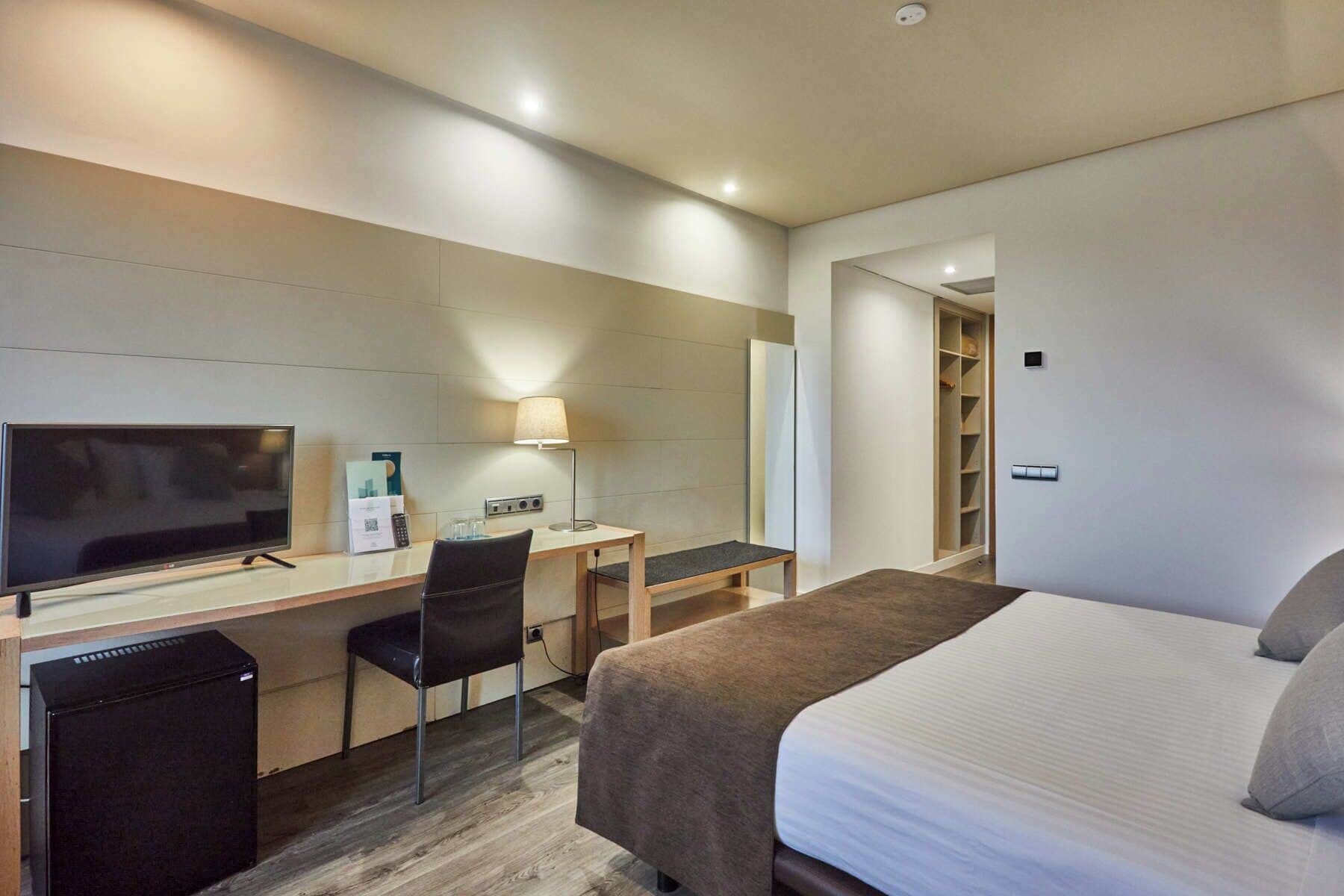 A hotel room with a bed, a desk with a chair, a television, a lamp, and a wardrobe.
