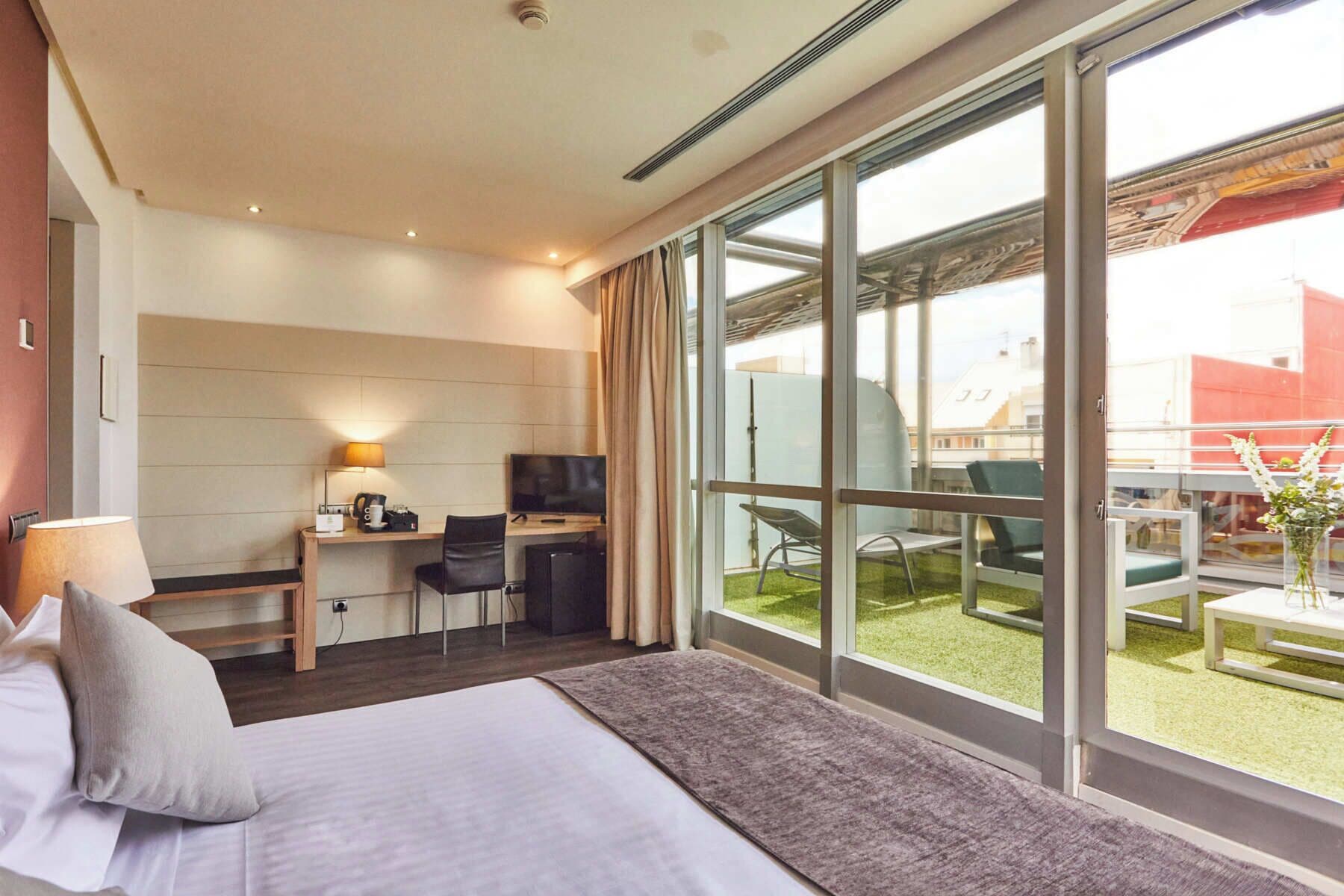 A hotel room with a bed, a desk with a TV, a chair, and a balcony with outdoor seating.