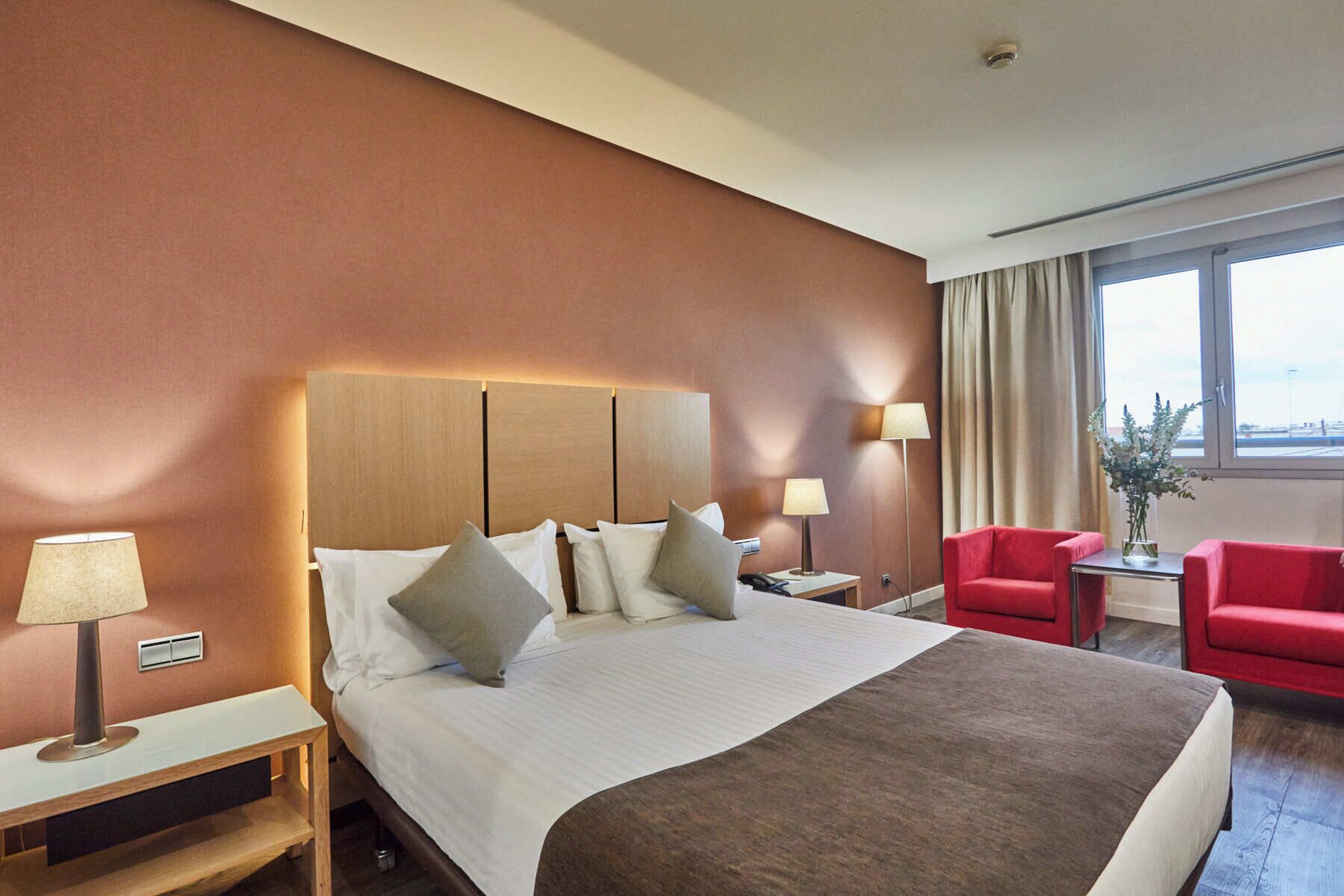 Une chambre d’hôtel avec un grand lit, deux tables de chevet avec des lampes, un fauteuil rouge et une vue sur l’extérieur par une fenêtre.
