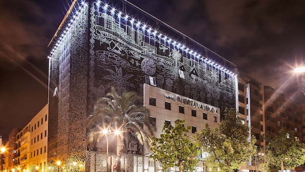 Een hotel met een grote, verlichte muurschildering op de gevel, waarop verschillende stadsgezichten en symbolen te zien zijn.