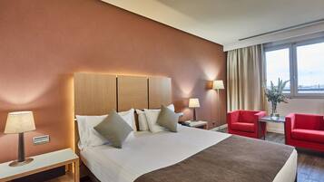 Habitación de hotel con una cama grande, mesitas de noche, un sillón rojo y vistas al exterior.