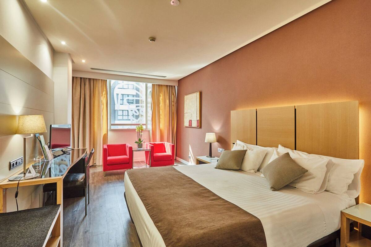 Een hotelkamer met een groot bed, een bureau met een computer, een stoel en een raam met gordijnen.