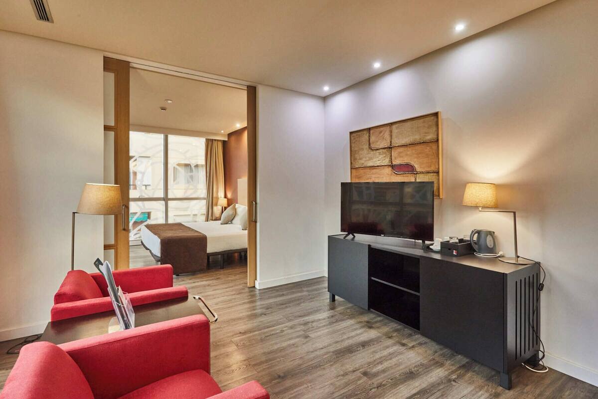 Een moderne hotelkamer met een houten vloer, een televisie op een zwart voetstuk, rode fauteuils en een groot raam met gordijnen.