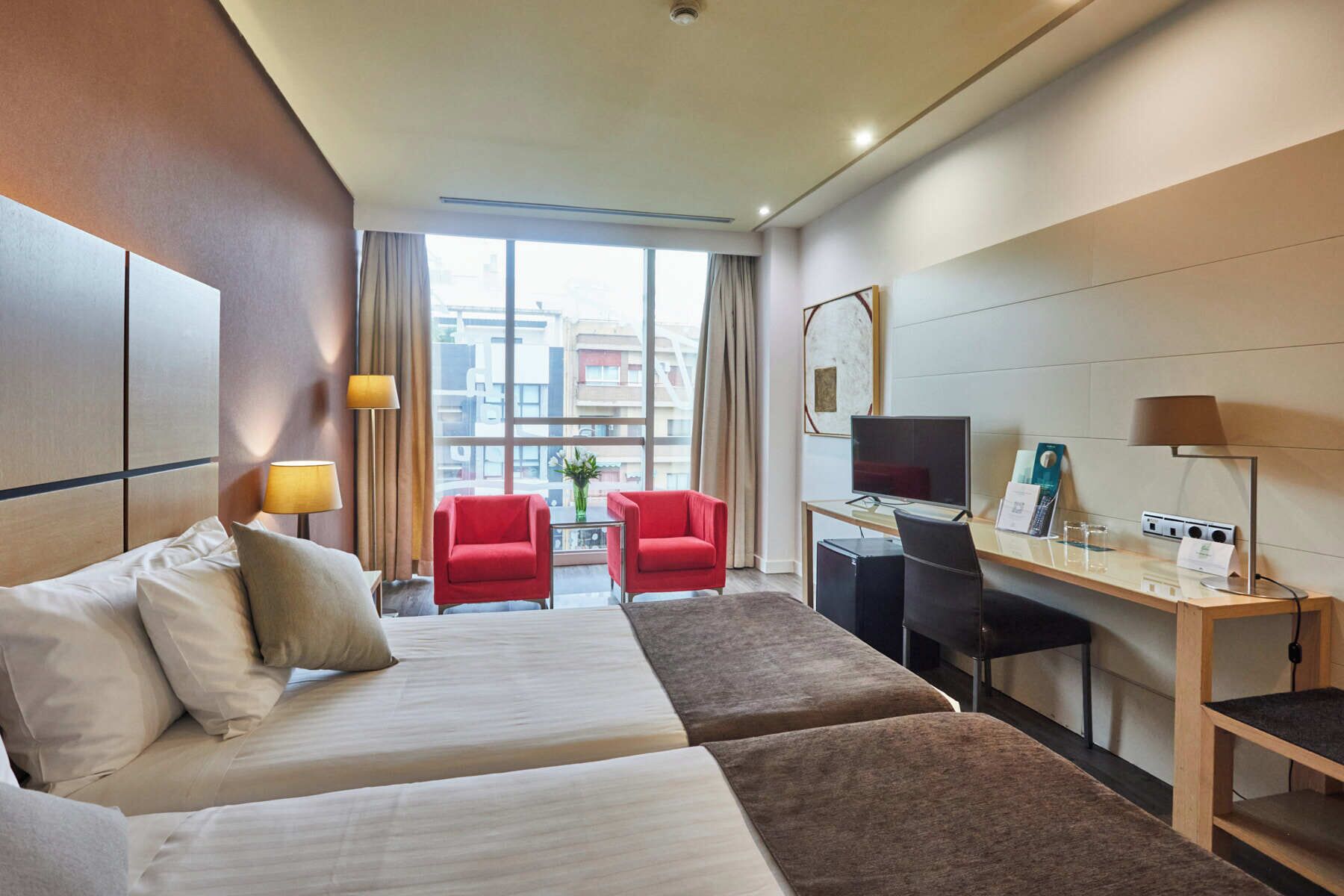 A hotel room with a bed, two red chairs, a desk with a computer, a TV, and a window with a city view.