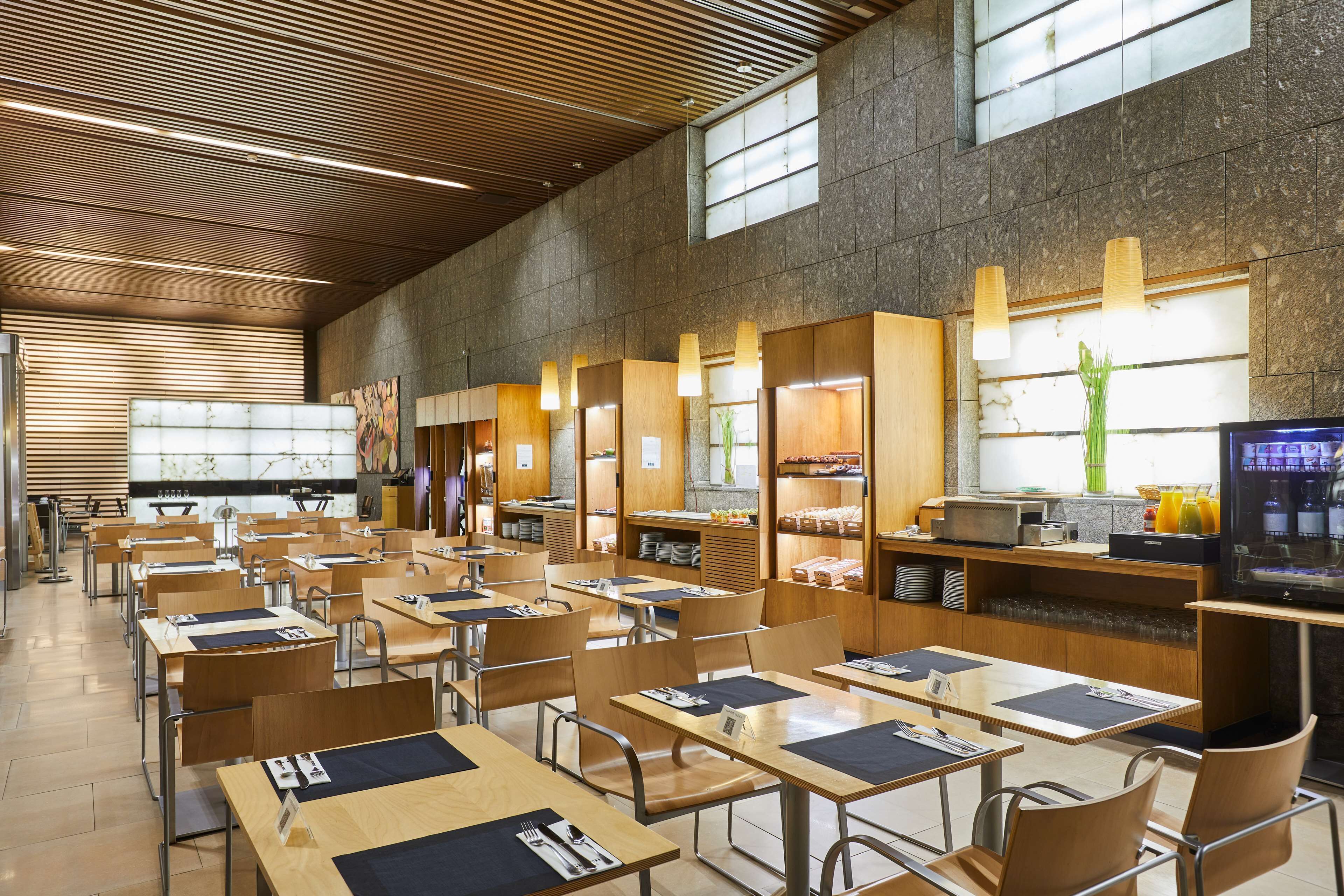 Una amplia cafetería con mesas y sillas largas, armarios de madera y un ventanal que proporciona luz natural.