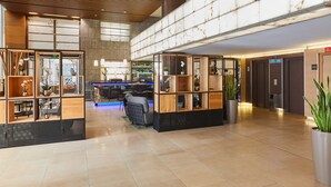 Een ruime lobby met een hoge plafond, houten schapenkasten met planten en bloemen, een zithoek met een bank en stoelen, en een bar in de achtergrond.