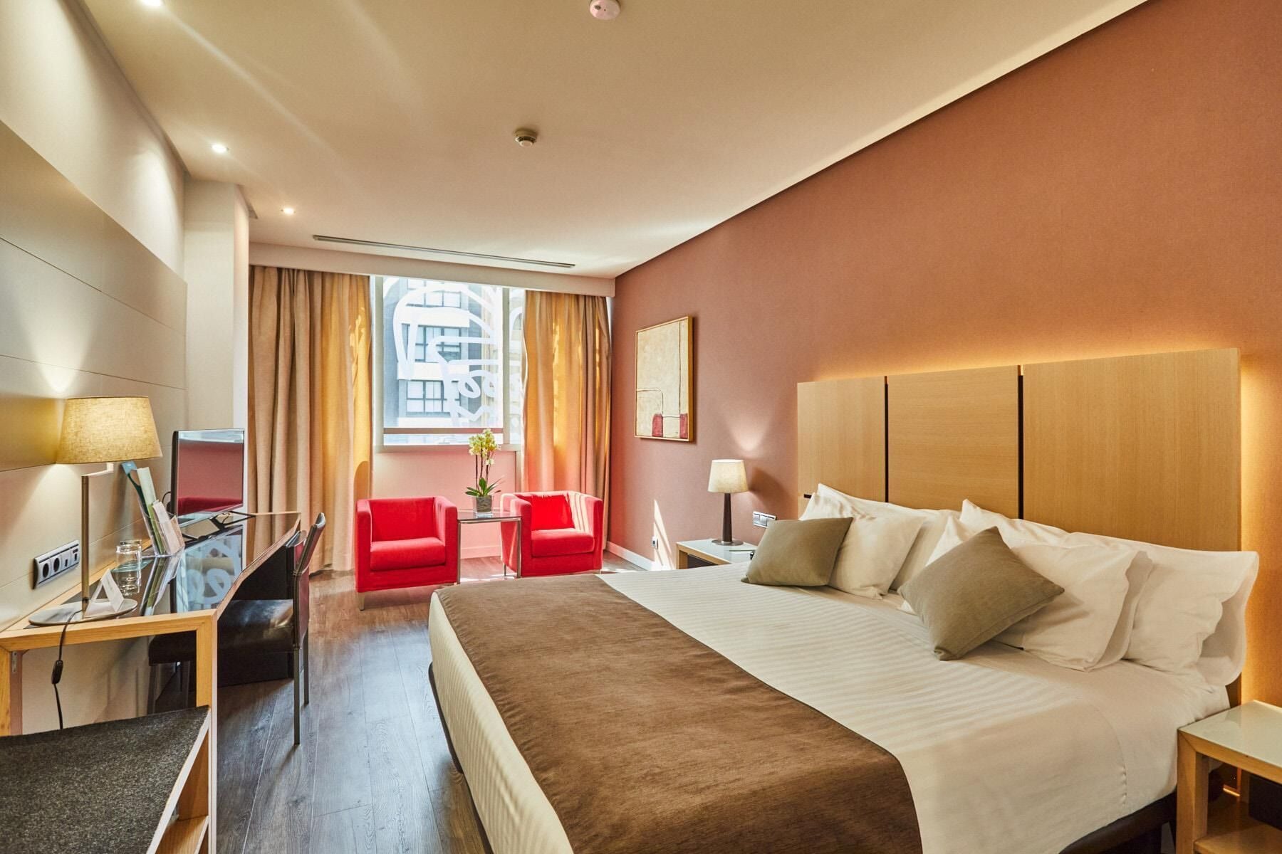 Ein Hotelzimmer mit einem großen Bett, einem Schreibtisch mit Lampe, einem Sessel und Blick auf die Stadt durch das Fenster.
